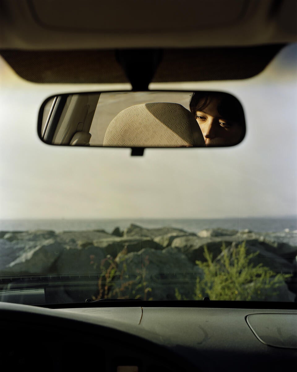 Photograph from inside of car looking out through the windshield to a rocky beach. The dashboard sits at the bottom of the image. There is a rearview mirror with a reflection of person face.