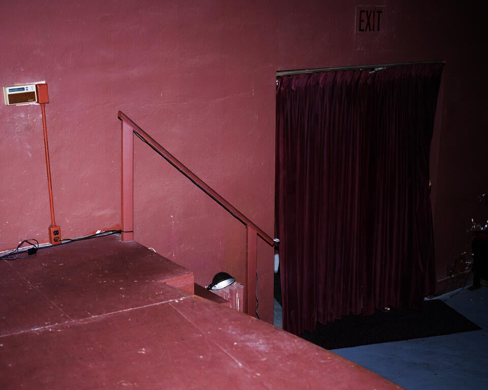 Photograph of a pink wall and stage. On the floor sits a doorway covered by a fushia velvet curtain. Behind the curtain an obscured person. Exit sign sits above the curtain  