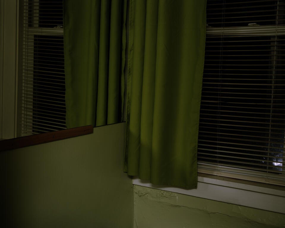 Photograph of a corner of a green room at night. On the right is a window with open blinds and an olive green curtain. Everything is reflected on the left through a mirror. 
