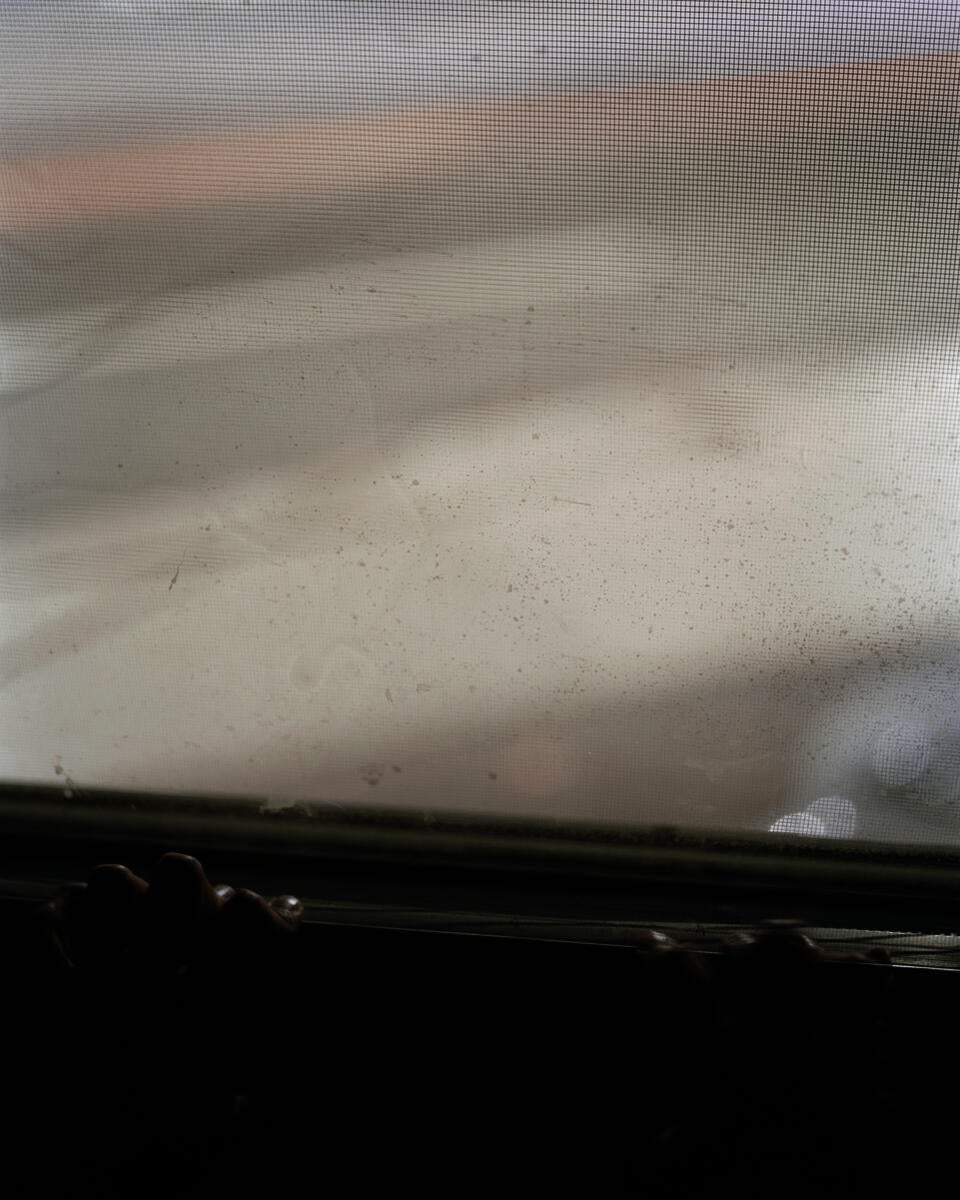 A long exposer photograph of a hands resting a on a white windowsill at night. The hands are mostly in shadow. Through the window is the gray street with long obscured car lights. 