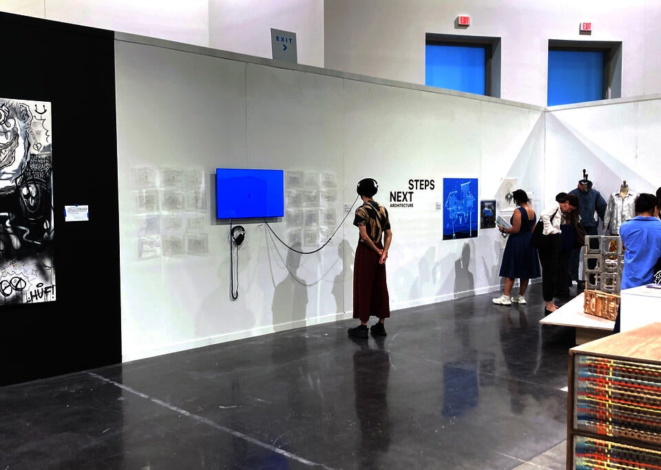 A single man reads the text on the wall next to a film installation. He is wearing headphones attached to the installation. People look at other art in the background.