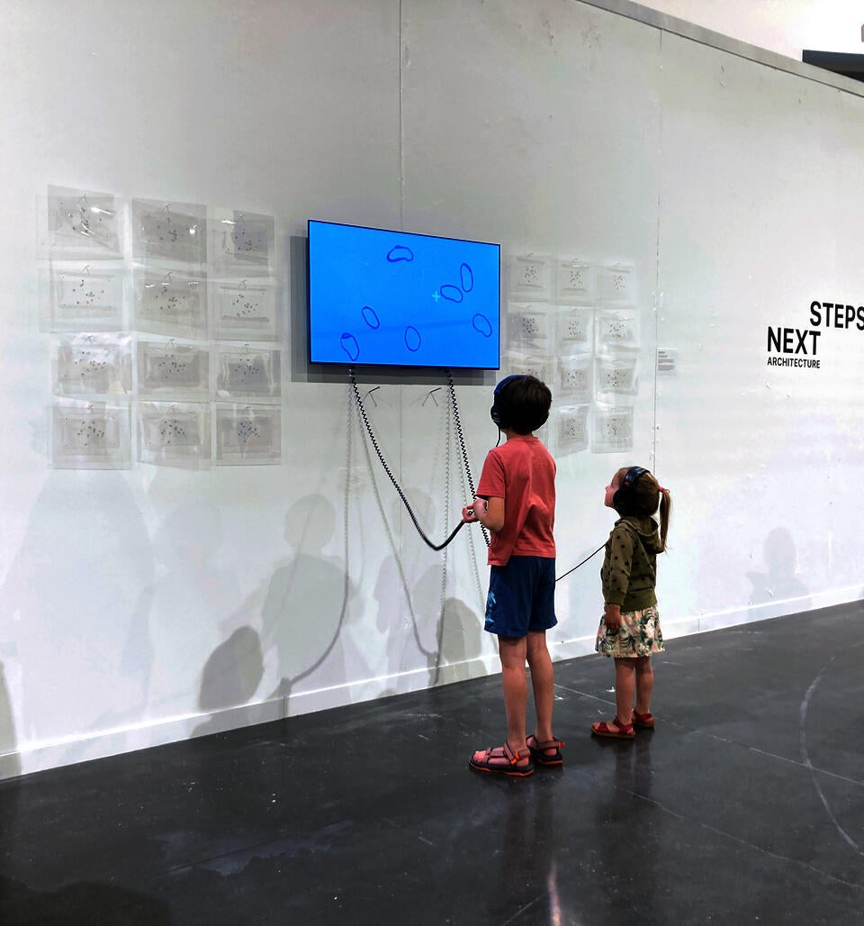 Two small children, both wearing headphones, gaze upward to watch a film installation. Shapes on the screen move against a backdrop of blue sky.
