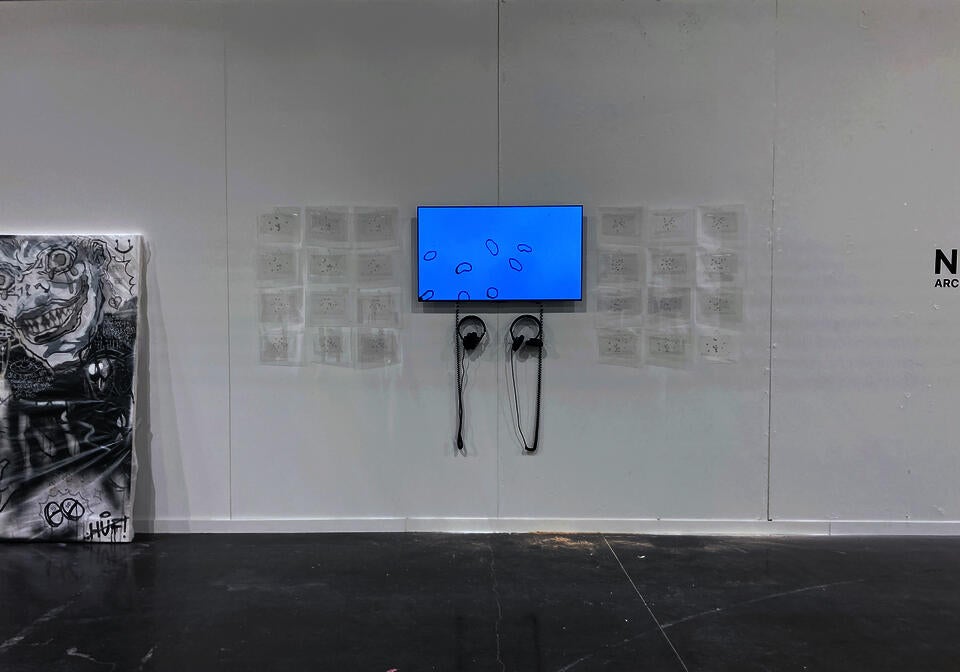 A TV is mounted on a white wall. On screen, shapes move against the background of a blue sky. Many small, transparent sheets hang neatly around the TV, two pairs of headphones hang beneath.