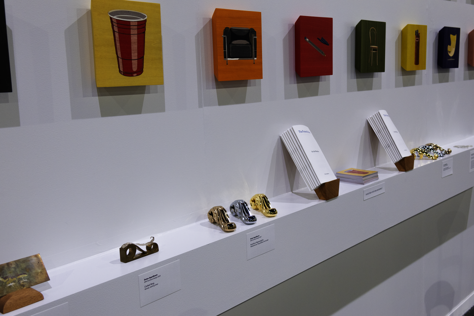 Left-side view of a shelf with Acanthus Paperweights, Corbel Ring, a decorative card scraper, booklets, and a stack of postcards. Above the shelf are colorful panels depicting chairs and ubiquitous products. 