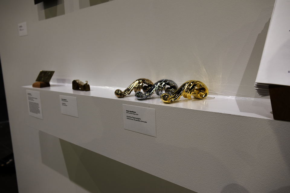 Right-side view of a white wall-hung shelf with Acanthus Paperweights, Corbel Ring, and a decorative card scraper. Title cards are posted on the front face of the shelf.