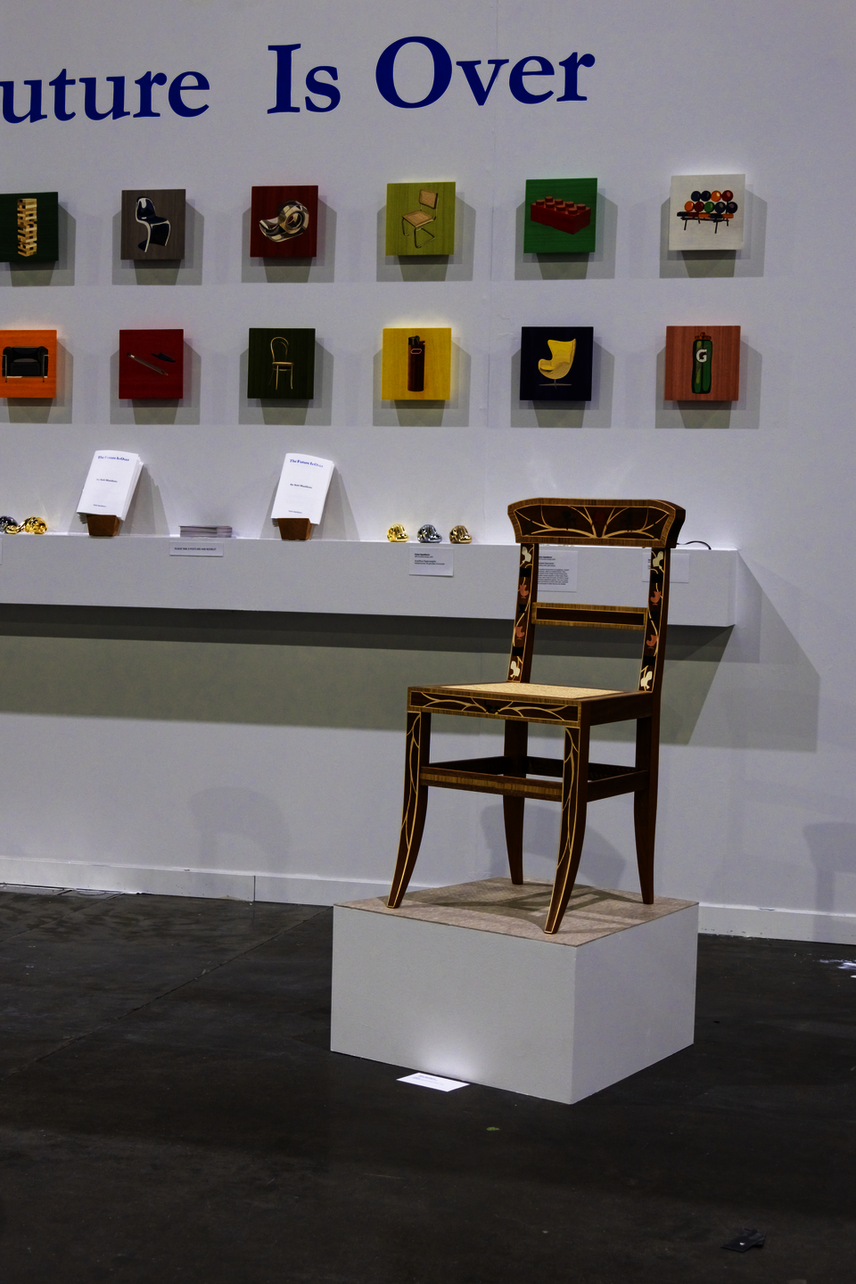 A chair with colorful inlaid depictions of chairs sits on a trapezoid-shaped pedestal with a linoleum top surface. Behind the chair is a wall with a shelf, colorful panels, and blue text.