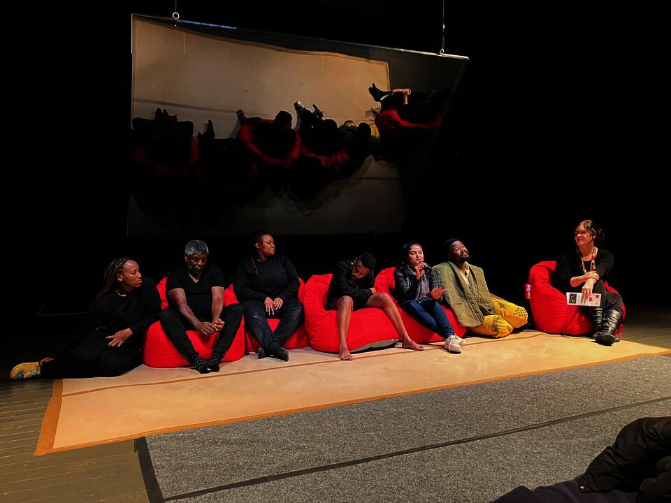 Individuals seated on red bean bags in front of the Pepper's Ghost Theatre Apparatus. Collaborators from William Kentridge's Centre for the Less good idea. Collaboraters are South African. Deanne Fernandes is present as well. 