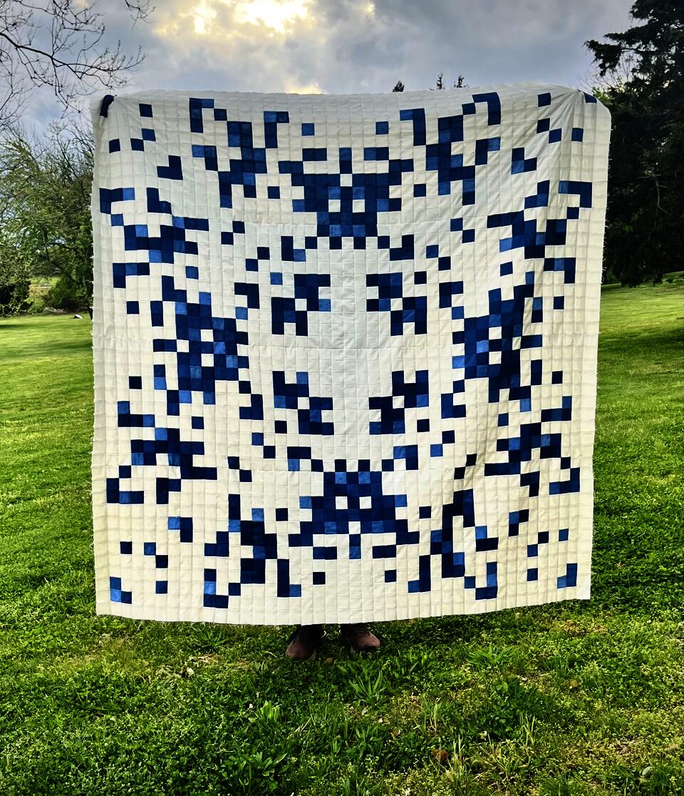 A large blue and white quilt is held up outside in a field of green grass at golden hour.