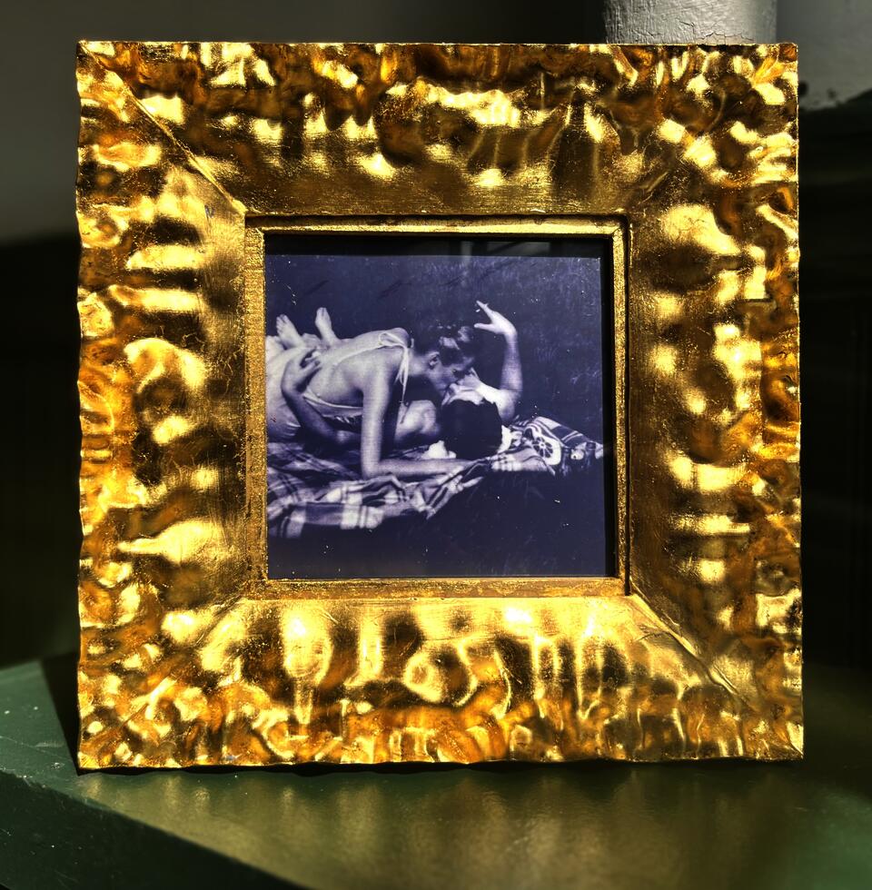 A golden frame holds a black-and-white vintage photo of two people kissing on a blanket in the park. 