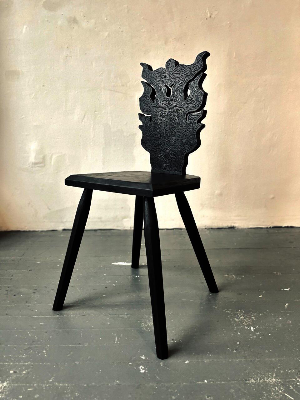 A black chair stands on a wooden floor. The base of the chair is simple, utilitarian and the back is intricately carved.