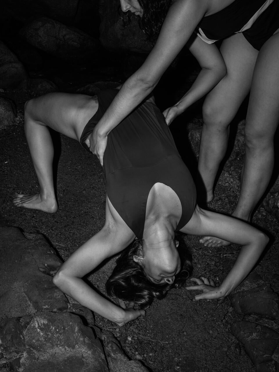 Black and white photograph of two people on the rocks of a beach, one person holding the other while she tries to make a complicated body posture
