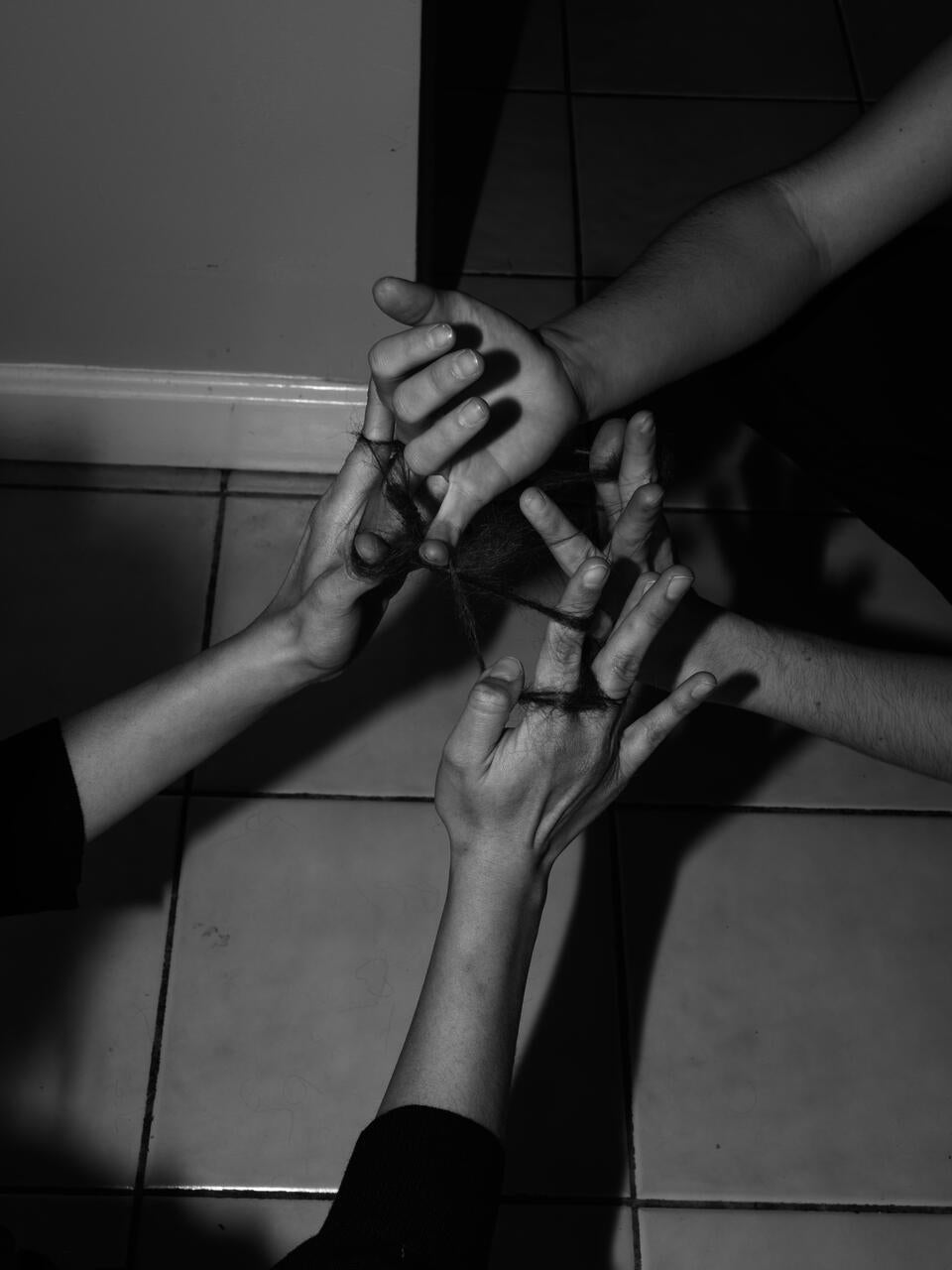 Black and white photograph of 4 hands playing cat's cradle in the kitchen of a house. Instead of a string they have human hair.