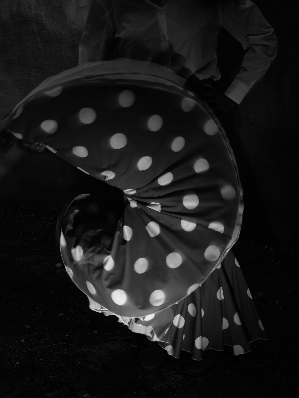 Black and white photograph of a person with a skirt full of white dots, which they are undulating with one hand for the picture 