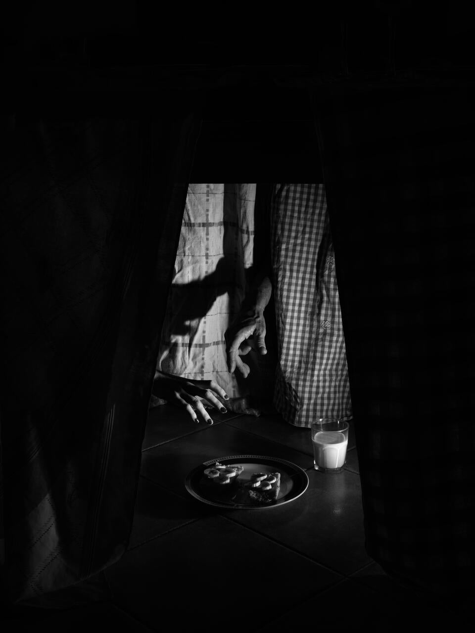 Black and white photograph of the bottom of a table, with 4 hands almost grabbing a plate of bread with banana and a glass of milk