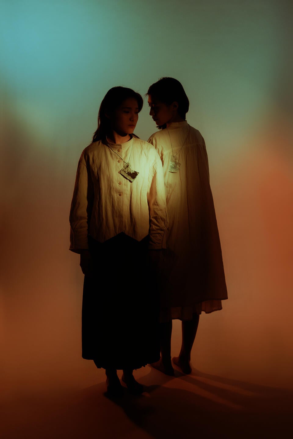 2 girls standing in the light of blue and orange mimicking sunrise, each person wearing a pendant. One face the front one face the back, looking at different directions. 