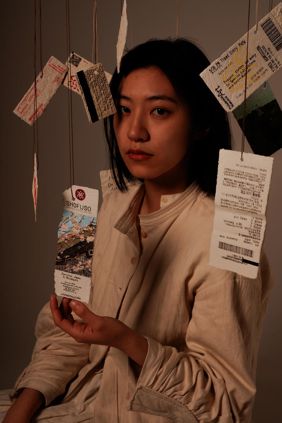 A girl wearing a cream dress surrounded by suspended tickets painted on paper mache. Her one hand is touching one ticket, eyes looking at the camera. 