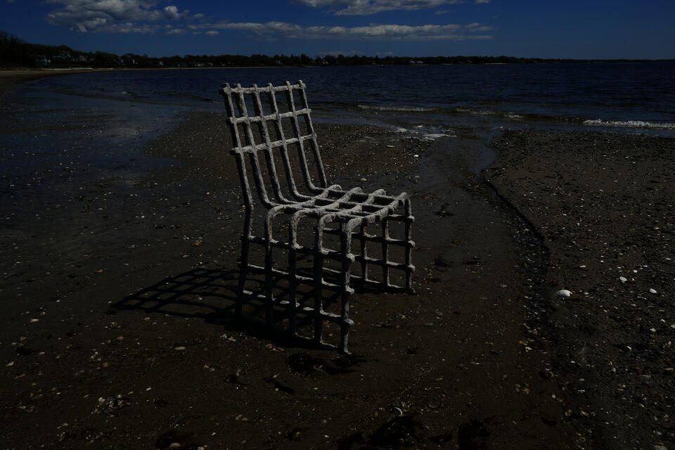 A gray and blue oyster and mussel biomaterial chair on a sandy, rocky beach by the water's edge. Houses and trees are visible across the water under a partly cloudy sky.