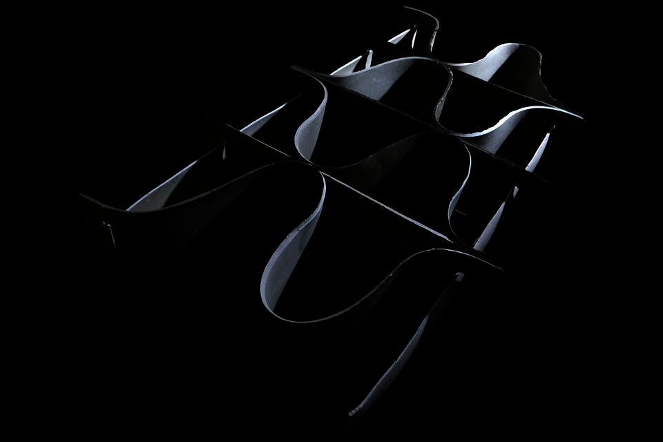 A shelf composed of seven thin, curved, ribbon-like forms intertwined and overlapping against a black background. The oyster biomaterial Shellf Life has a reflective, silver-toned surface that catches the light for a beautiful furniture design.