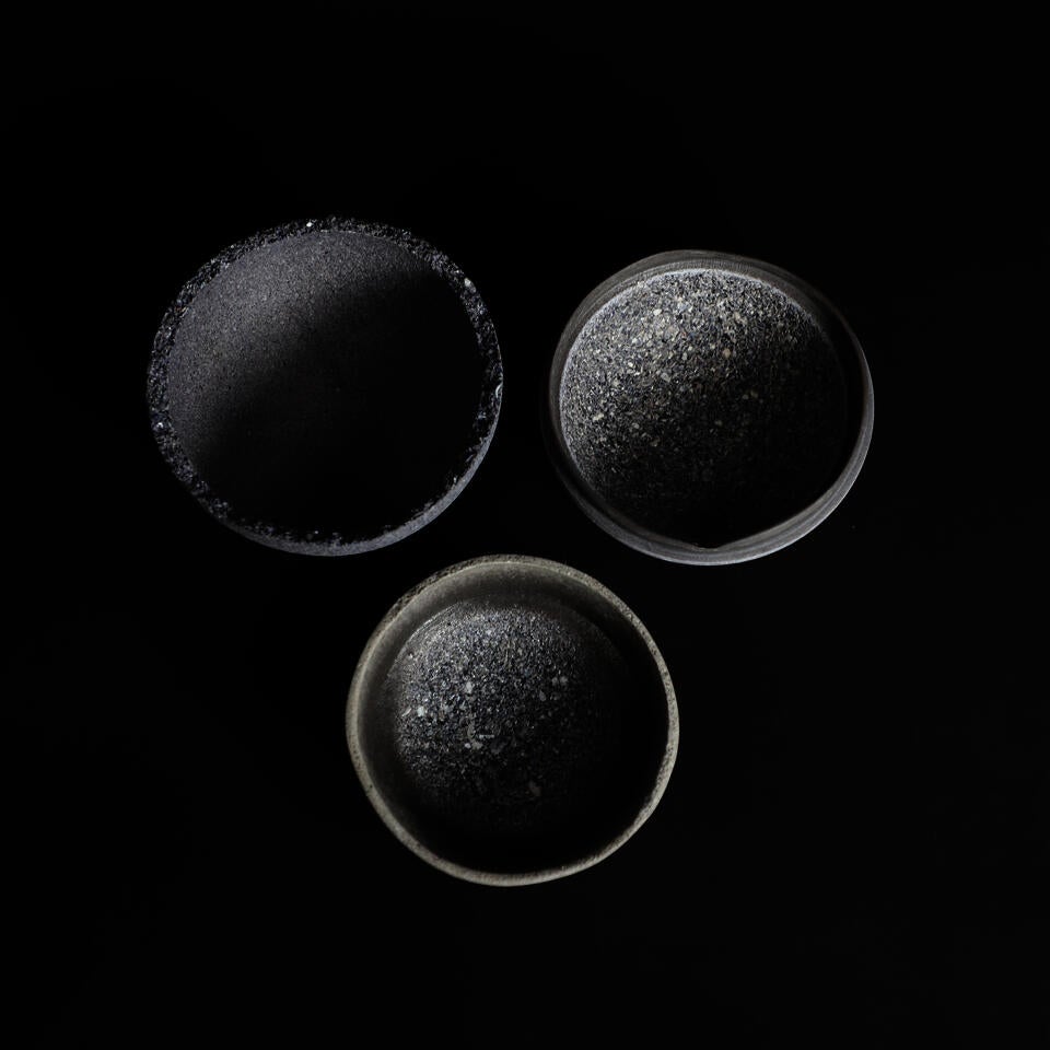 Three shallow, round bowls made from mussel shells, arranged in descending size on a black background. The bowls have textured, dark blue exterior surfaces and smooth, light gray and blue interior finishes.