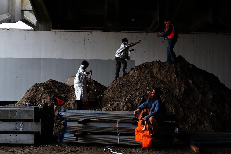 Photograph of four people wearing the following garments:100% Recycled Denim Suit and Jacket, two Tyvek Jackets, cork chaps, and an orange nylon vest stuffed with recycled denim pulp, in a construction site under an overpass.