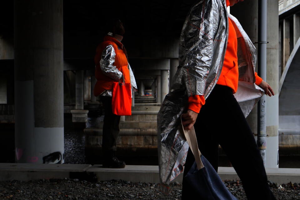 Photograph of two people wearing the Tyvek Trench and Tyvek Coat layered with a Shoddy Vest under an overpass, holding bags.