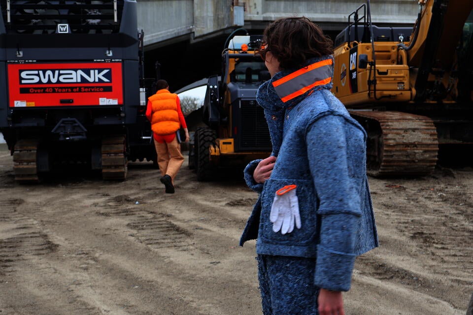 Photograph of two people, one in the foreground wearing the Denim Insulation Suit, the other person is walking away, wearing the orange Shoddy Vest and Cork Chaps.