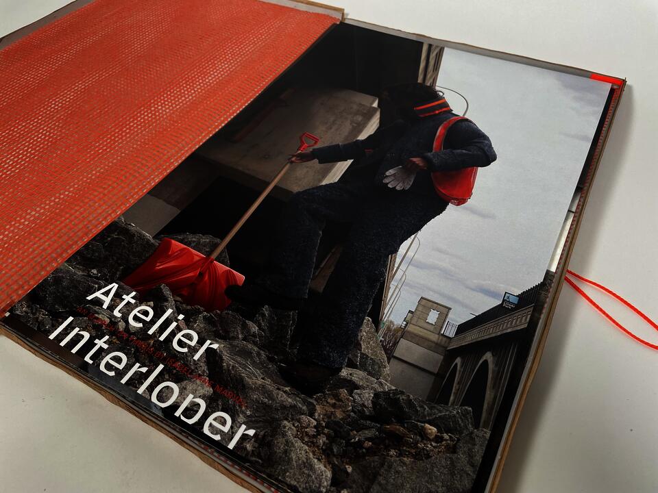Photograph of the collection's lookbook opened to the cover page, a photograph of a person in the denim insulation suit climbing the mountain of rubble with an orange shovel. Title of the page reads "Atelier Interloper"