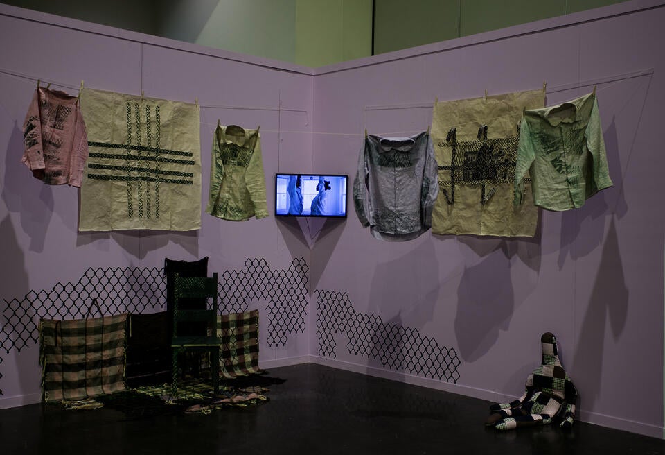 Image of printed clothes hanging from clothesline, video performance, woodcut prints, carved chair, woven fabrics, and stuffed doll