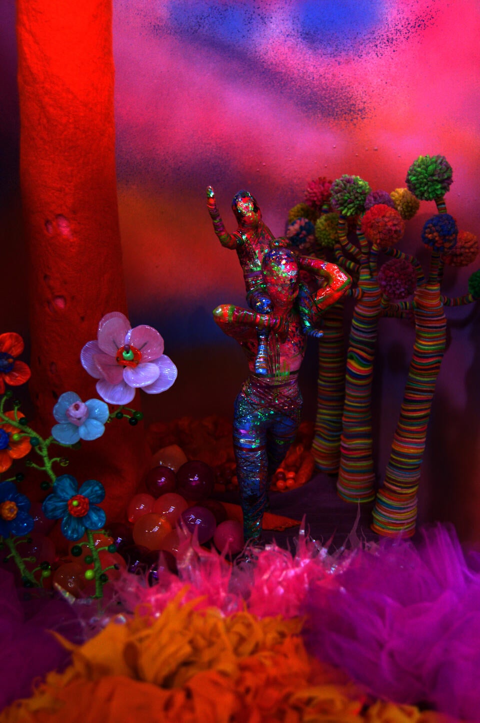 A colorful, glittering child sits atop the shoulders of a similarly colored adult as they walk through a fantasy forest with striped wool trees and large glass flowers