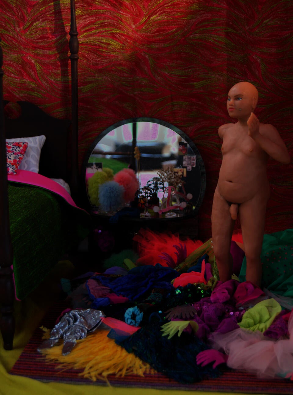 A sculpted nude figure stands in a bedroom, the floor covered with elaborate, brightly-colored clothes. A mirrored vanity has ruffled headdresses on glass wigstands, a post bed is covered with a green and pink silk duvet.