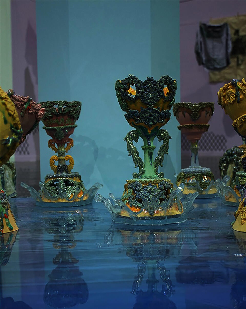 Close-up of intricately decorated the Holy grail with dripping glazed surfaces and covered with pastel colors, standing on a glossy blue reflective surface which is water.