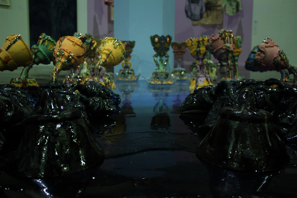 Foreground features dark, glossy ceramic forms with a rugged texture to express water, leading to pastel-colored, ornate vessels in the background on a reflective surface. 