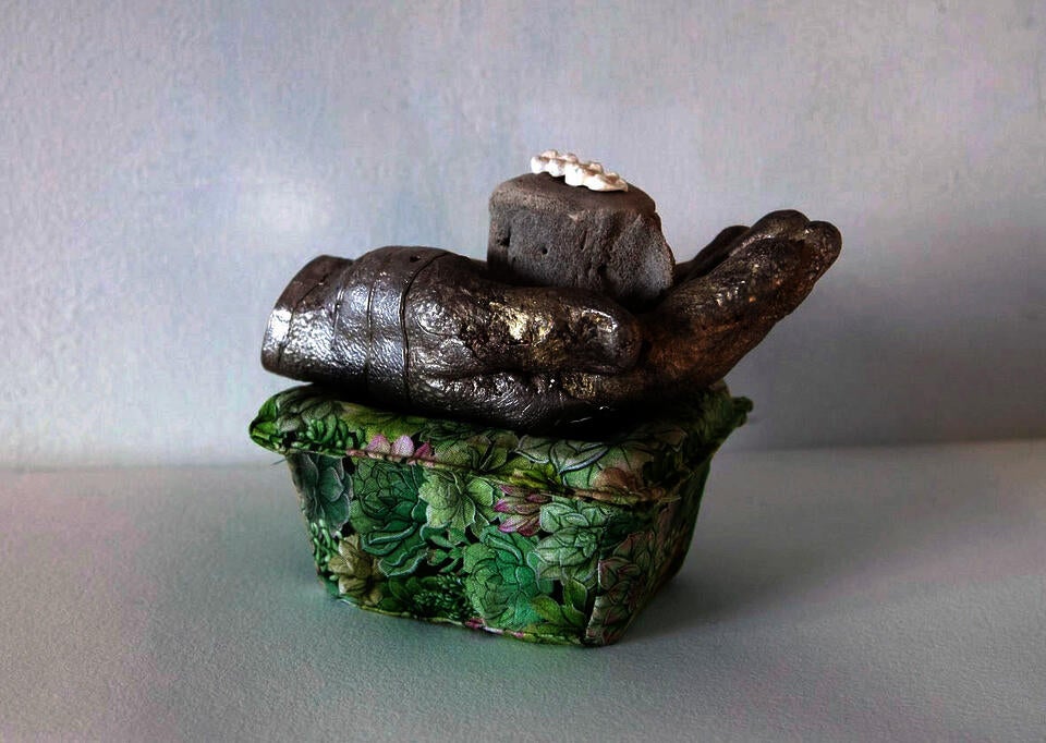 a stack of objects; a white mold of three molar teeth, an ocean animal vertebrae, a pewter hand that is crushing a blackberry plastic container that is covered in green leaf patterned fabric