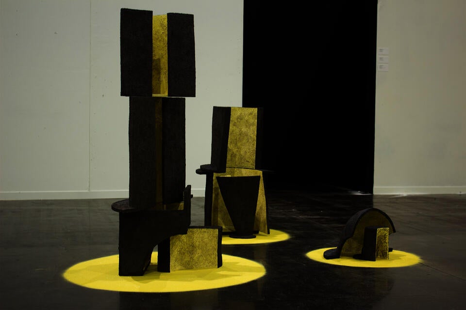 A collection of textured, geometric ceramic sculptures in various shapes, predominantly yellow with black accents, displayed on yellow circular bases.