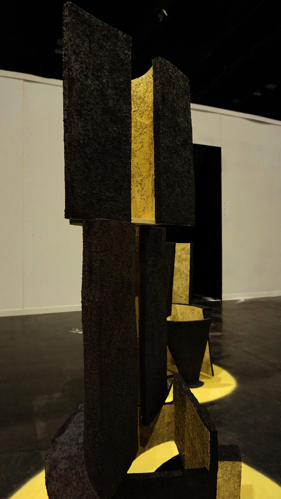Close-up of a textured, geometric ceramic sculpture with vertical panels in black and yellow, displayed on a yellow circular base. The background shows additional sculptures with similar designs.