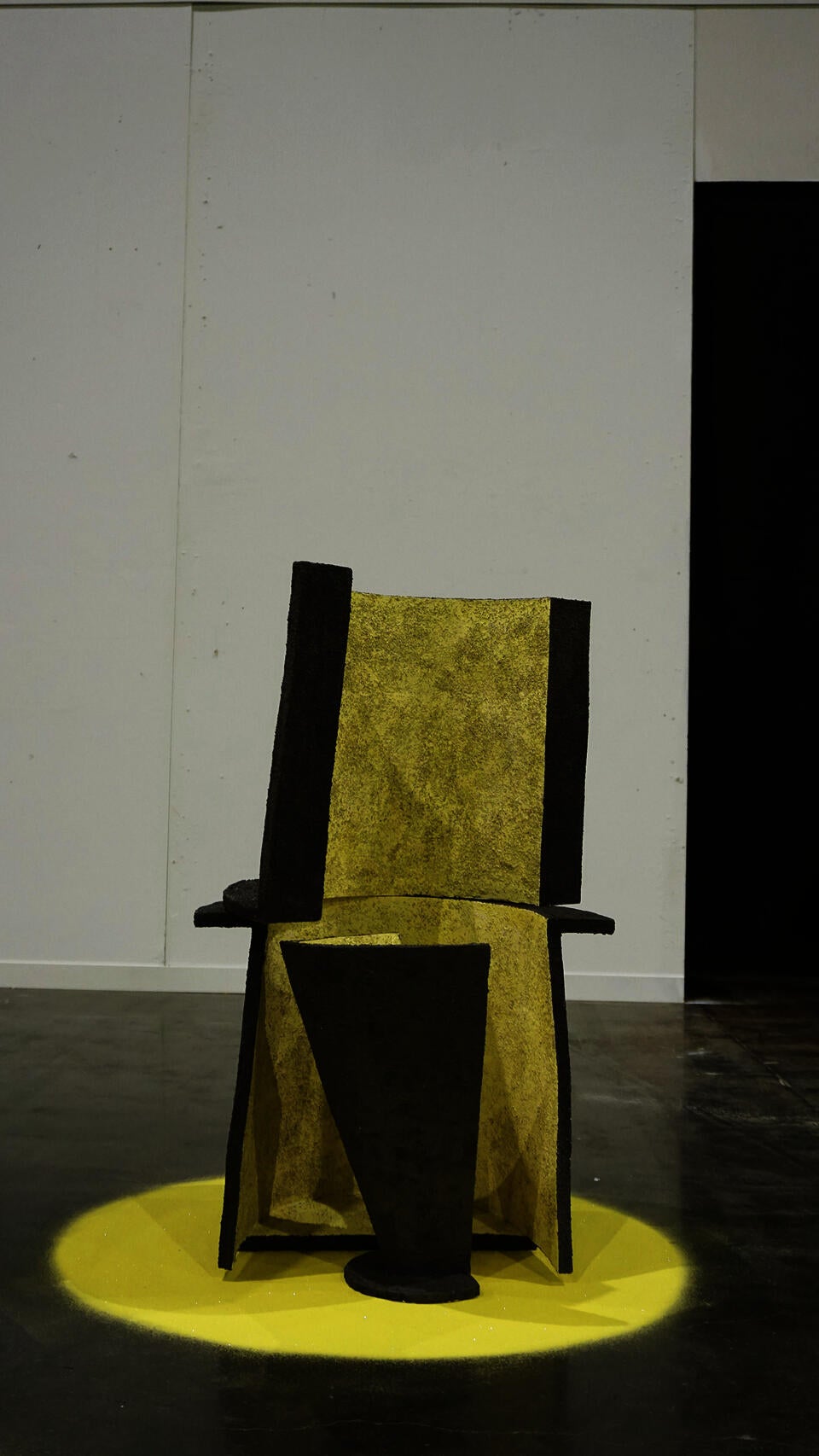 A textured, geometric ceramic chair with a tall back and angular seat, predominantly yellow with black accents, is placed on a yellow circular base in a minimalist gallery space.