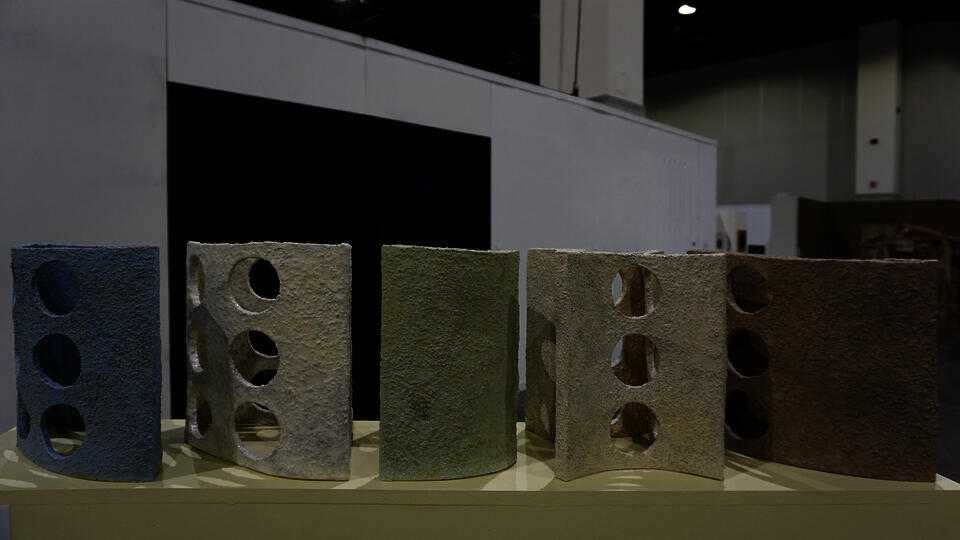 A row of textured, crescent-shaped ceramic sculptures with circular cutouts, arranged in gradient of earth tones and pastels. The foreground shows beige, transitioning to green and blue in the background.
