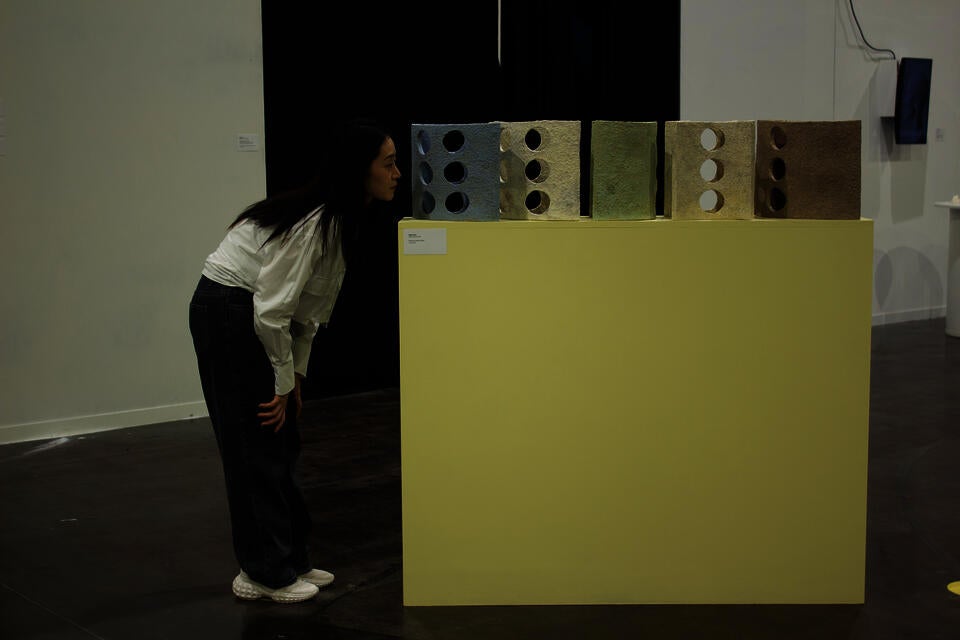 A woman in a white blouse and dark jeans leans forward to closely examine a series of textured, crescent-shaped ceramic sculptures with circular cutouts, displayed on a light yellow pedestal.