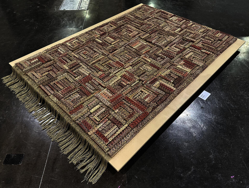 3D-printed ceramic (Egyptian paste) piece with intricate patterns in shades of red, beige, and brown. The first row is made of yarn with hanging threads. The piece is displayed on a wooden platform, placed on a polished concrete floor.
