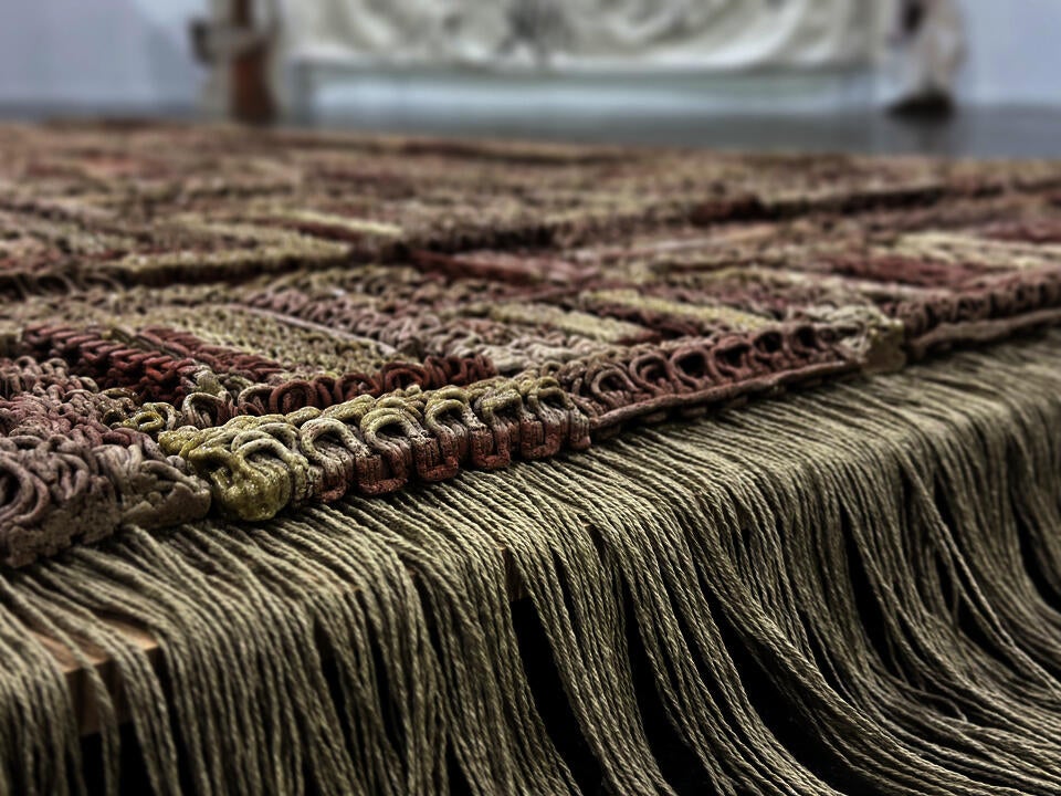 3D-printed ceramic (Egyptian paste) piece with intricate patterns in shades of red, beige, and brown. The first row is made of yarn with hanging threads. The piece is displayed on a wooden platform, placed on a polished concrete floor.