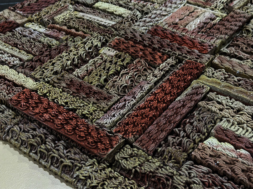 3D-printed ceramic (Egyptian paste) piece with intricate patterns in shades of red, beige, and brown. The first row is made of yarn with hanging threads. The piece is displayed on a wooden platform, placed on a polished concrete floor.
