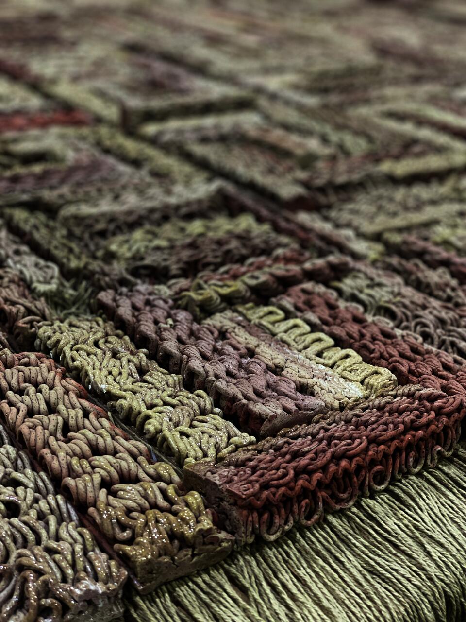 3D-printed ceramic (Egyptian paste) piece with intricate patterns in shades of red, beige, and brown. The first row is made of yarn with hanging threads. The piece is displayed on a wooden platform, placed on a polished concrete floor.