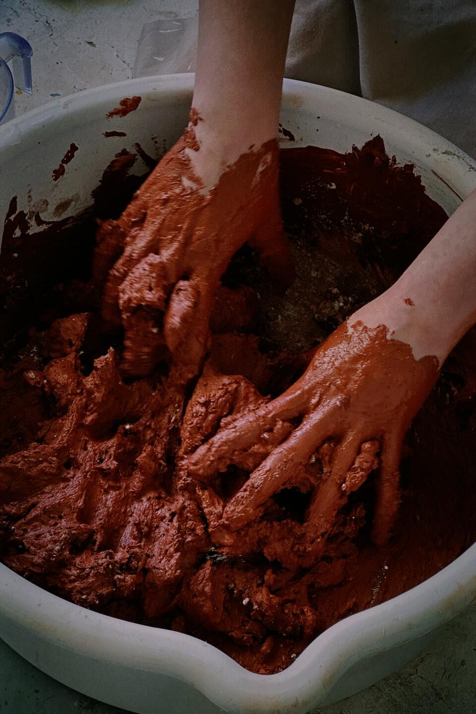 Hands mixing reddish Egyptian Paste in a white bowl, creating a textured, earthy mixture. The scene captures the tactile and immersive process of preparing ceramic material.