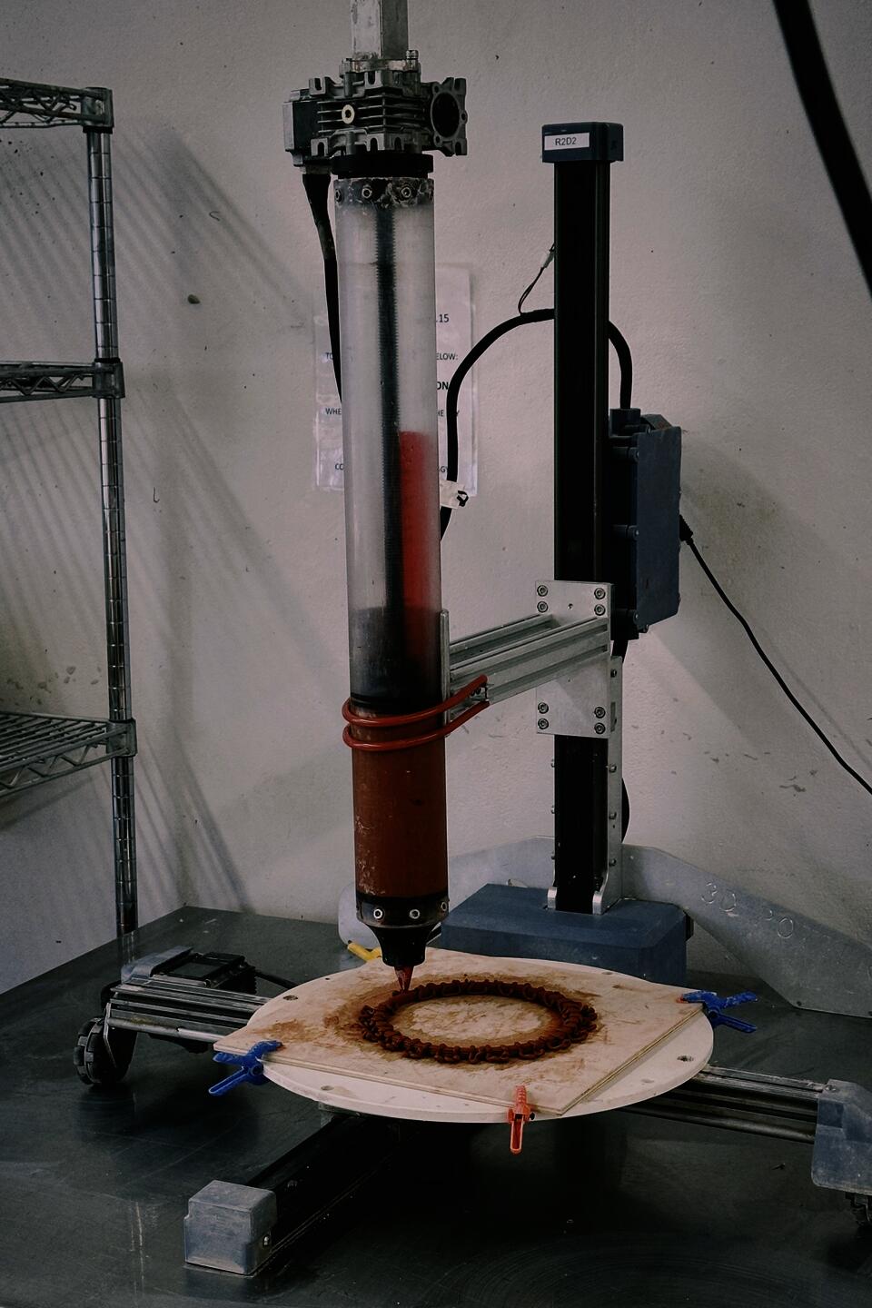 3D printer extruding a circular clay pattern on a rotating platform, with the printer labeled 'R2D2' in a workshop setting.
