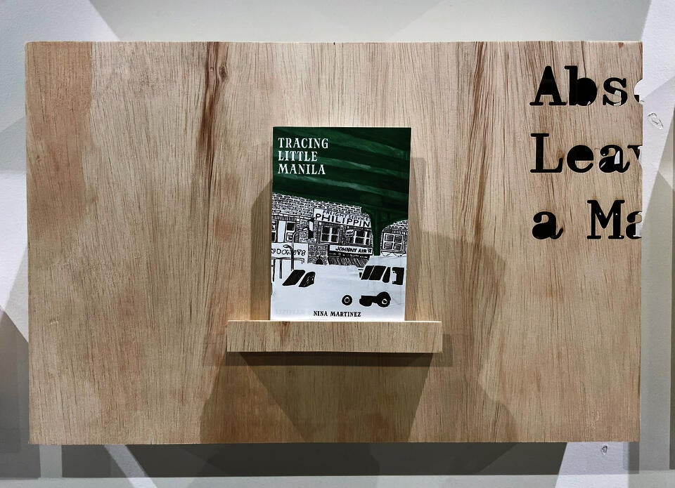 A closeup of the Tracing Little Manila zine, sitting on a small shelf on a plywood display. The text "Absence Leaves a Mark," the title of my thesis, is laser cut into the corner.