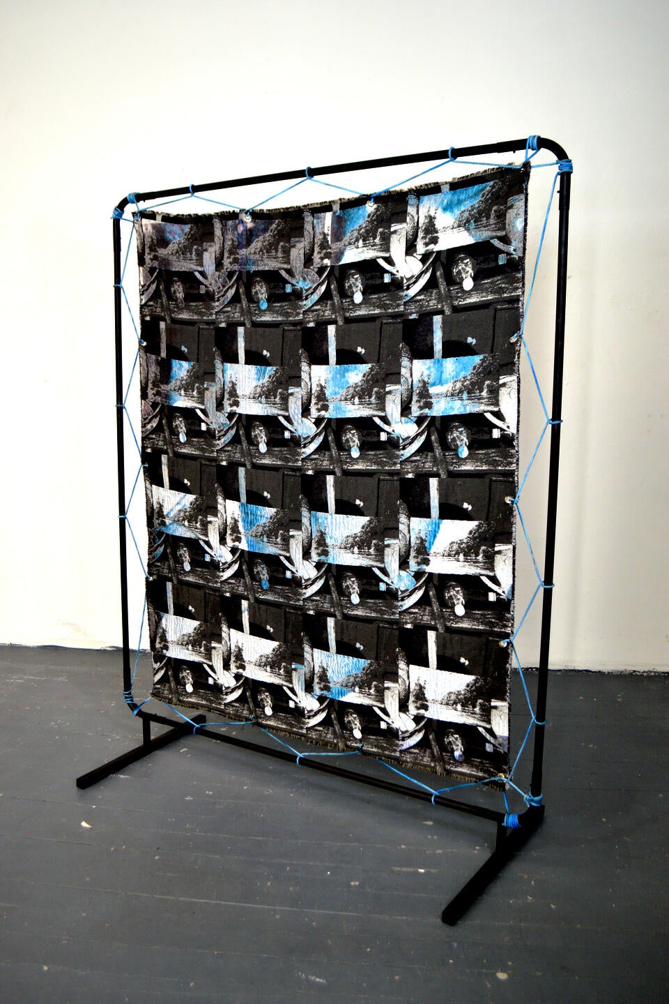 Large black steel frame standing in a studio facing a blank wall with a black and white photographic weaving depicting an image repeat. The weaving is stretched using a blue rope.