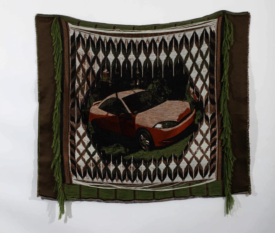 A medium sized square weaving depicting an orange car in the middle with a black and white pattern around it hanging on a white wall.