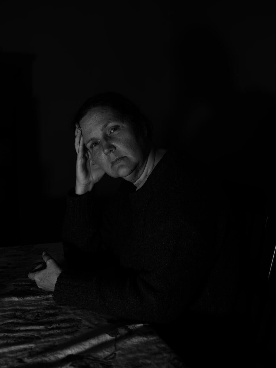 A woman in her mid-50s sat in a dimly lit dining room. Her shadow projected onto the wall behind her. 