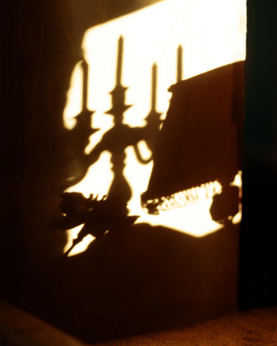 An interior space with a wall covered in sunlight from a nearby window. The wall features a reflection of a table lamp, a single flower, and candelabra.