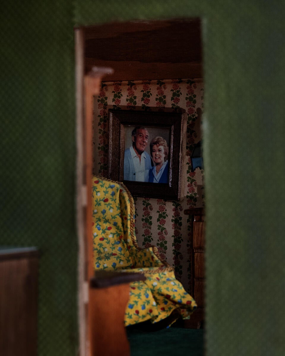  smeftah_text smeftah_text 100% 10 R2  An interior space, scaled at the perspective of looking through a doorway to another adjacent room. In the frame of the doorway is a portrait of an older couple on the wall, along with, a yellow floral chair, floral wallpaper and green carpet.      	 An interior space, scaled at the perspective of looking through a doorway to another adjacent room. In the frame of the doorway is a portrait of an older couple on the wall, along with, a yellow floral chair, floral wallpa